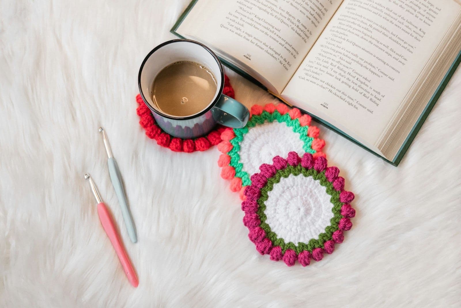 Round crochet coasters with bobble edges beside a mug of coffee, two crochet hooks, and an open book (inspiration photo by another maker, not made from this pattern)