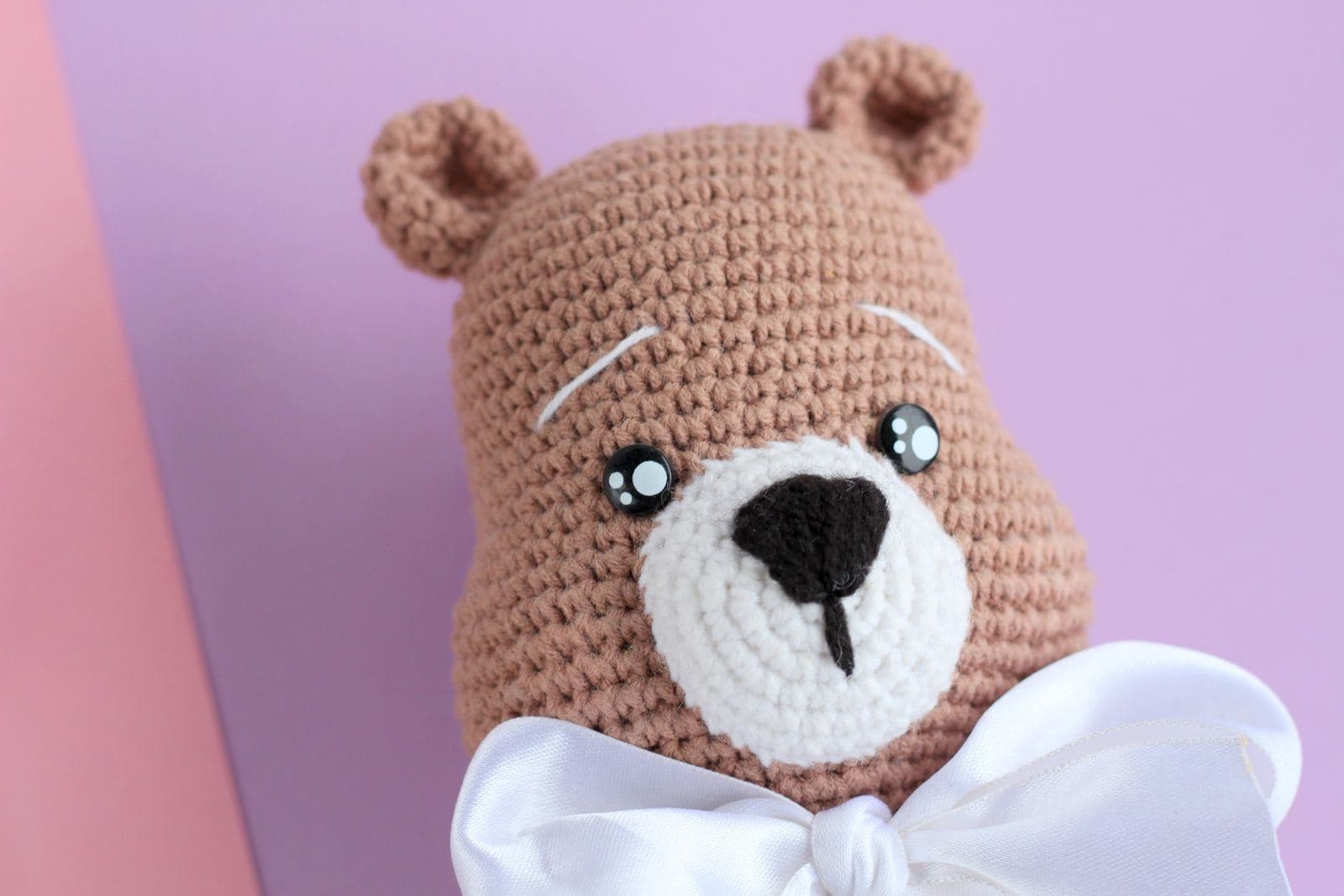 A tan amigurumi crochet bear with a cream muzzle and a white satin bow on a lavender background