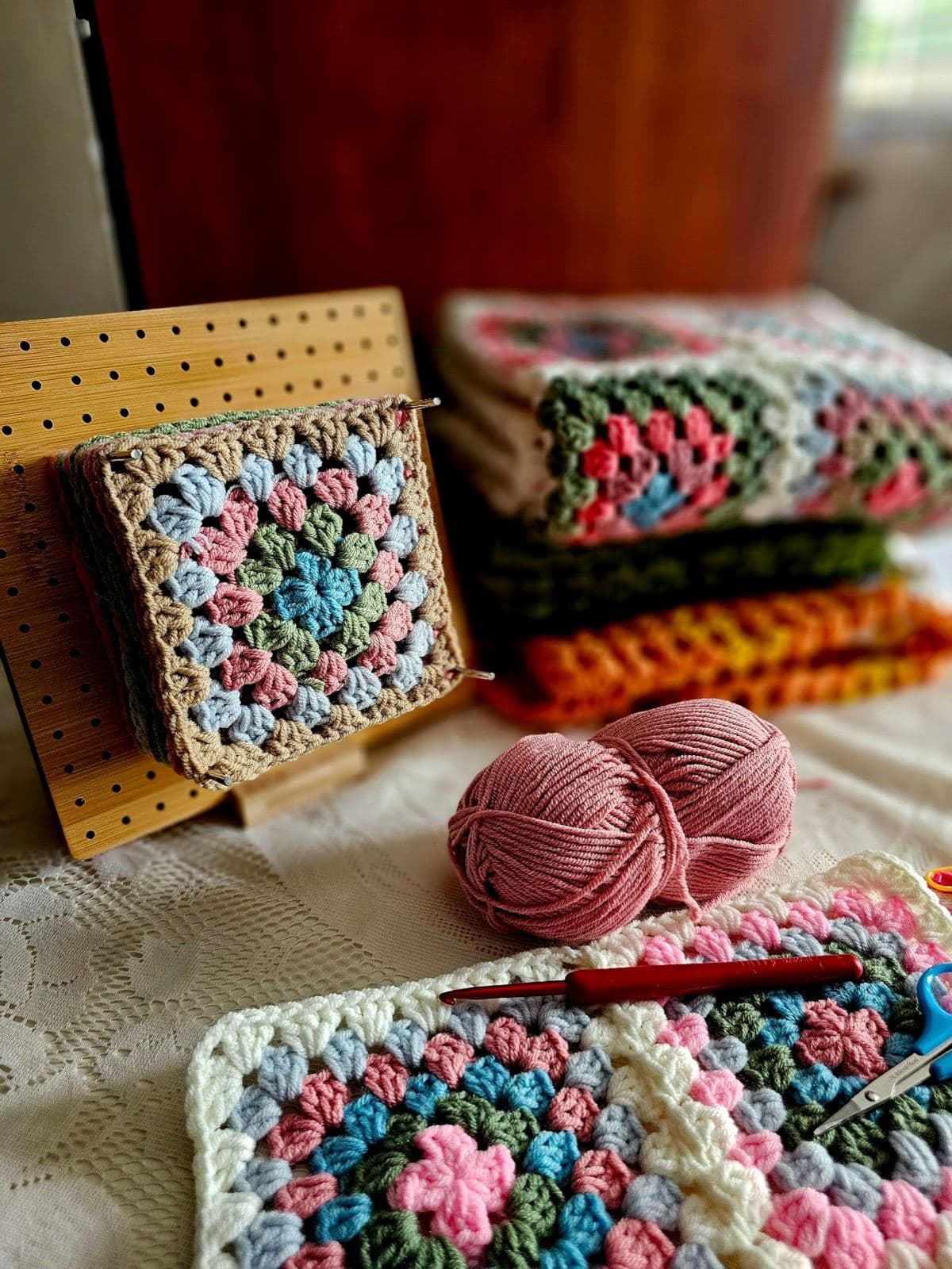 Granny square blankets, a ball of pink yarn, and a crochet hook on a bed