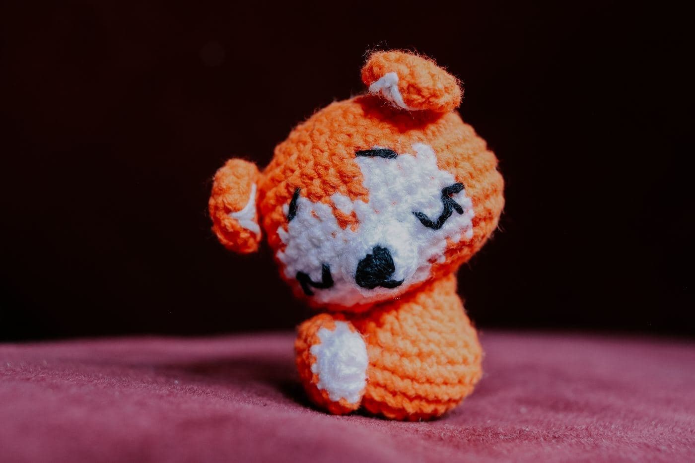 A small orange and white amigurumi animal with a round crocheted head on a pink cushion (inspiration photo by another maker)