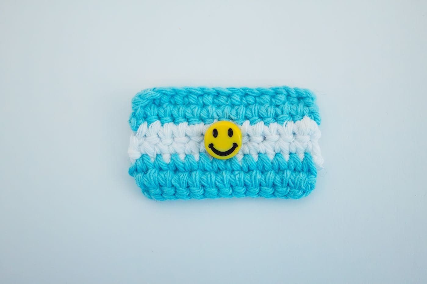 A small blue and white striped crochet pouch with a yellow smiley-face button on a pale blue background (inspiration photo by another maker)