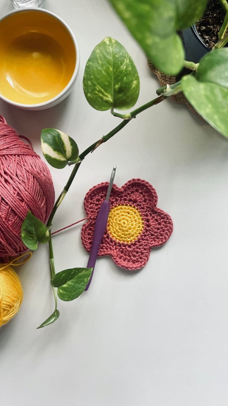 A red crochet flower with five rounded petals and a yellow center beside a purple crochet hook, pink yarn, and a plant (inspiration photo by another maker)