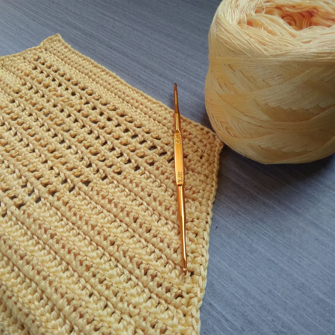 Rows of pale yellow crochet fabric with a gold crochet hook and a ball of matching yarn (inspiration photo by another maker)