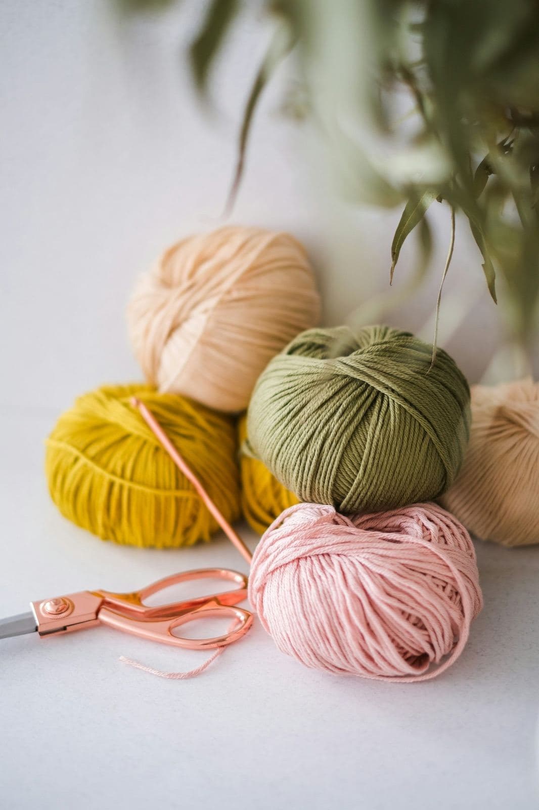 Balls of peach, green, mustard, and pink yarn with rose-gold scissors and a crochet hook